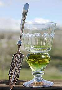 Een glas gevuld met een natuurlijk gekleurde wervel. Daarnaast is er een absintlepel.