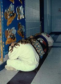 Uczniowie biorą udział w ćwiczeniach na wypadek tornada, ustawiają się wzdłuż wewnętrznej ściany i zakrywają głowy.