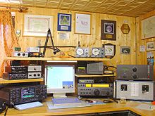 Un ejemplo de estación de radioaficionado con cuatro transceptores, amplificadores y un ordenador para el registro y para los modos digitales. En la pared hay ejemplos de varios premios, certificados y una tarjeta de informe de recepción (tarjeta QSL) de una estación de aficionado extranjera.  