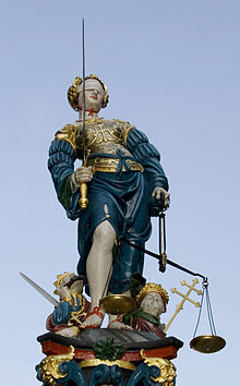 The allegorical representation of justice as Justitia, common in Western culture. She usually has three attributes: the scales (weighing, arbitrating, private law), sword (condemning, criminal law), and a bandage before the eyes (regardless of the person).