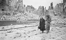 A British soldier helps an old woman after the conquest of Caen. The ruins of the city can be seen in the background.