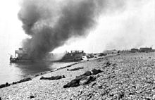Destruída a embarcação de desembarque em chamas com mortos canadenses na praia. Uma colocação de arma de concreto à direita cobre toda a praia. A praia íngreme pode ser claramente vista.