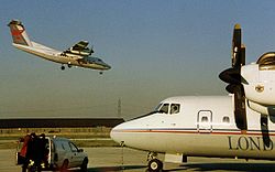 de Havilland Canada Dash 7 извършва стръмен подход към LCY от запад, докато друг DHC-7 на London City Airways се подготвя да отлети за Амстердам.