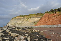 Falha normal na costa de Somerset, Inglaterra. À esquerda, a rocha cinza-azul mais jovem é Triássica tardia a Jurássica precoce. À direita, a rocha vermelha é o arenito Triássico mais antigo. Os estratos da rocha vermelha se deformam quando se move para cima. A linha da falha percorre a praia, mostrada pela rocha/areia dividida.