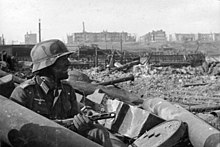 Wehrmacht Feldwebel Wilhelm Traub beväpnad med en PPSh-41 skannar av utsikten över Stalingrad mitt i en förstörd stad hösten 1942. Den förstörda staden är i dag känd som Gagarin Park.  