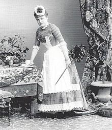 Maid in hall, slightly bent standing at table with feather duster, studio photo.