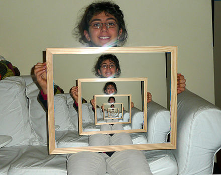 Infinite reflection as an example of recursion: The person sits facing a larger wall mirror with the mirror held up. The following mirror image contains itself as a part.