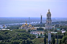 The cave monastery and the Mother-Home statue situated on the hills of the western bank of the Dnieper, behind it the Dnieper and its shallow eastern shore.