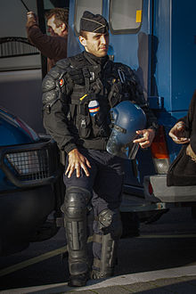 Un gendarme con armadura que protege sus extremidades y su cuerpo y que lleva un casco antidisturbios de SPECTRA con visera.  